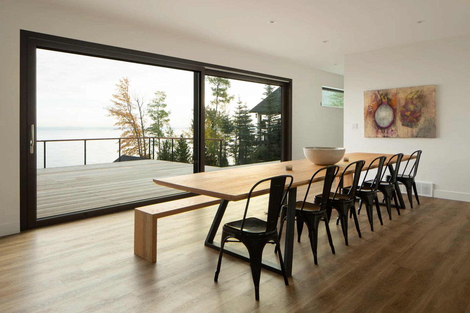 Long dining table beside a full-height black-frame sliding glass wall with the water horizon beyond